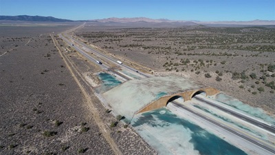 An aerial view of a large wildlife crossing over a split highway set into a largely empty landscape with some hills. The highway is split into two roads, so the crossing has two bridge portions. There are a few large green shrubs throughout the landscape, which is otherwise gray-brown soil. A deep blue sky is at the top of the image.