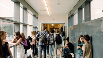 Students gather in a brightly lit hallway between classes, some walking with backpacks and others leaning against green walls, chatting with friends.