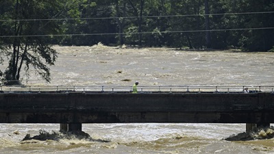 A man in a yellow vest walks across a bridge above high brown floodwaters.