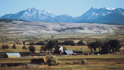 Mountain peaks rise behind ranch buildings, a hay meadow, and a haystack.